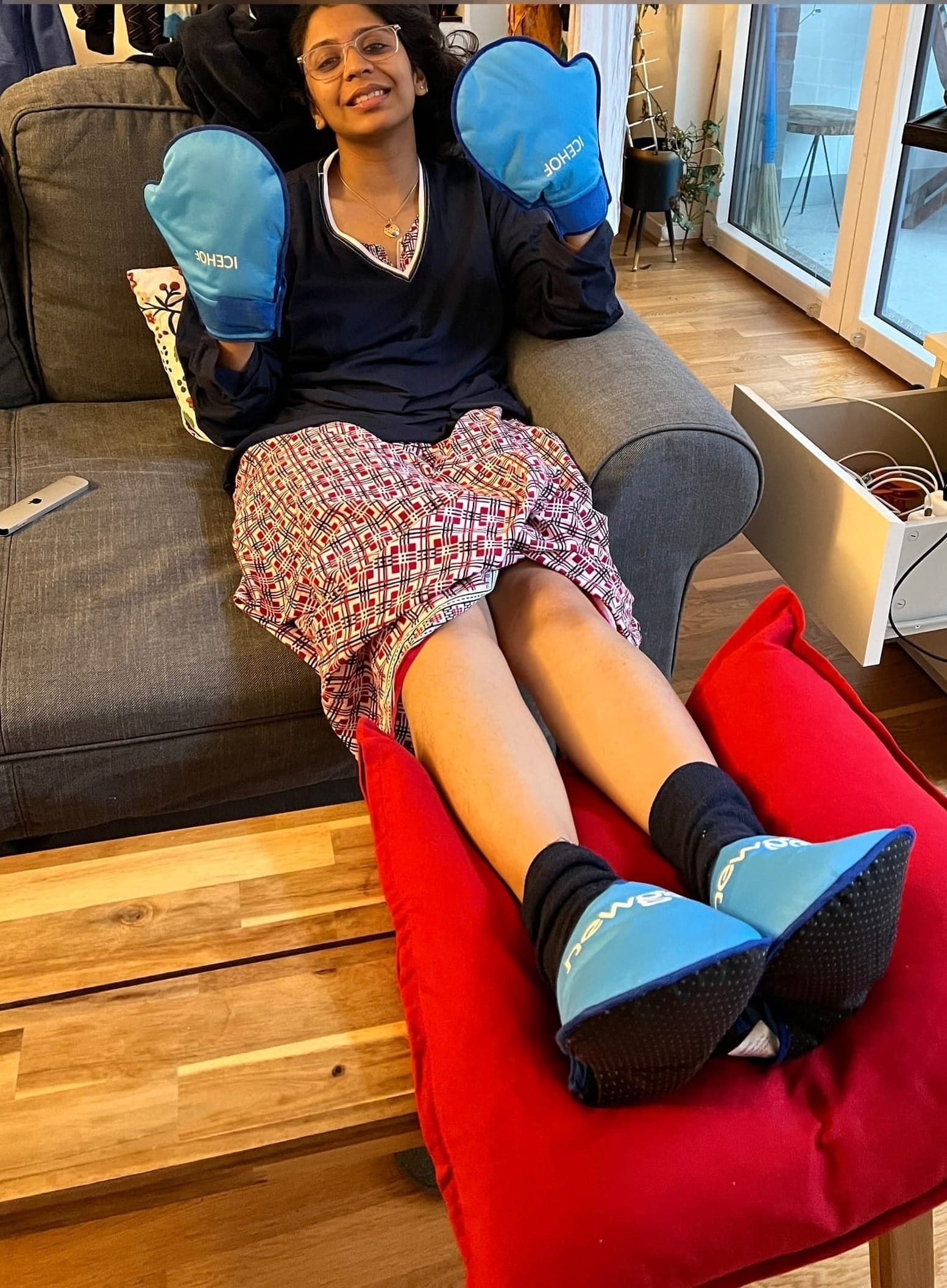 Swathi seated on a grey sofa with her feet outstretched onto a table opposite to her. She's wearing heat packs on her feet and hands.