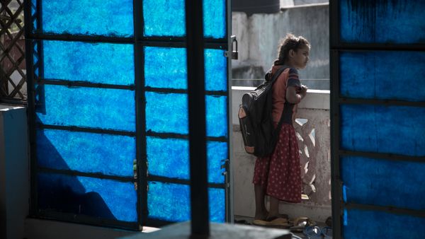 A young Indian girl aged 7 or 8 looks out on to the street from her balcony. The photo has been taken from inside the house 