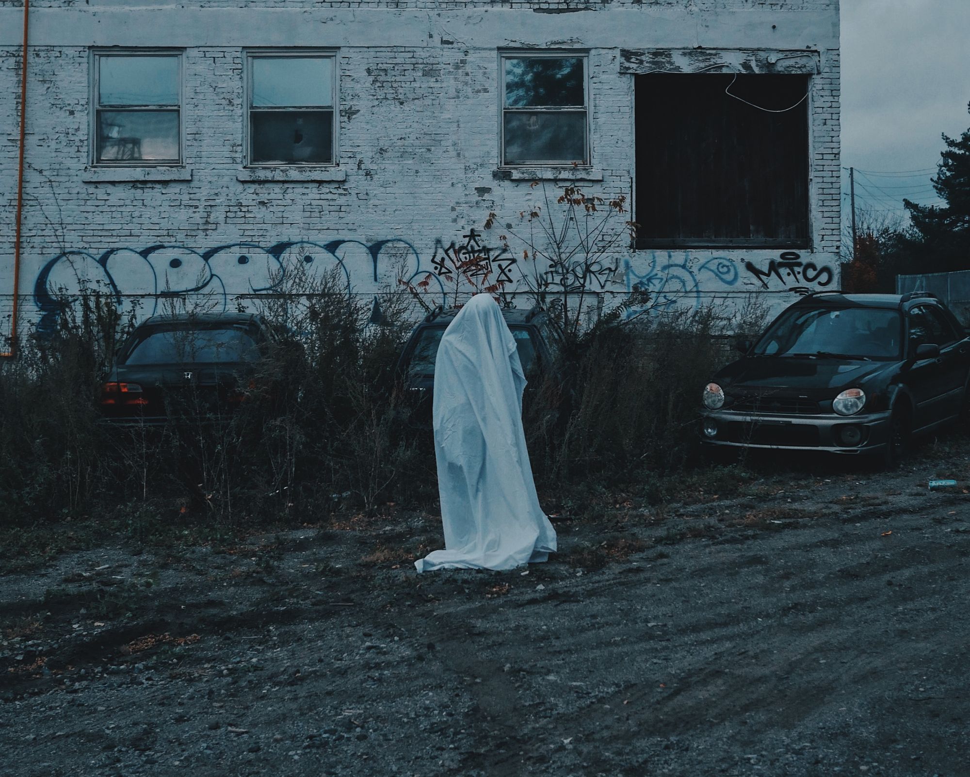 A person is standing and they're covered from head to toe in a white bedspread in an abandoned car park.